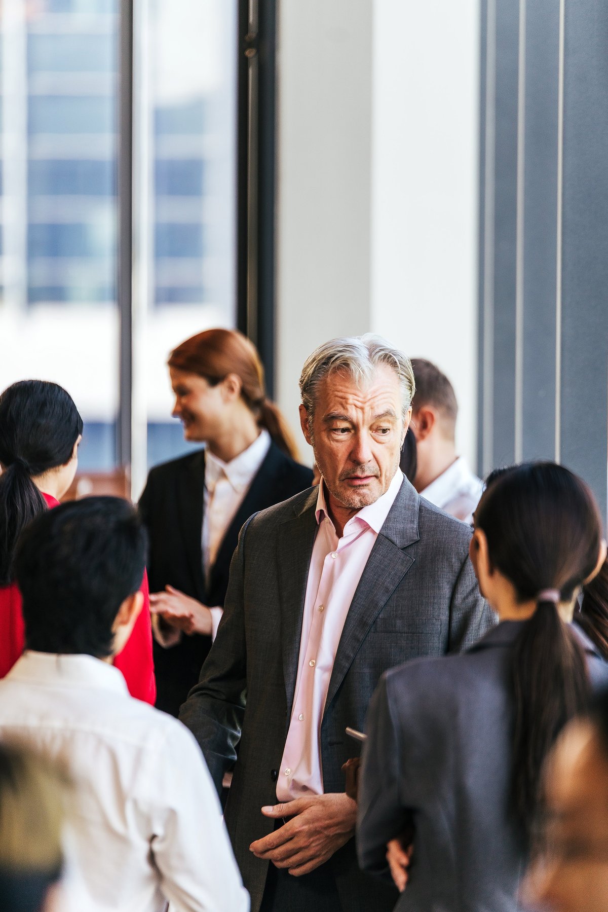 Professionals in suits network and converse warmly at a corporate event near large windows.