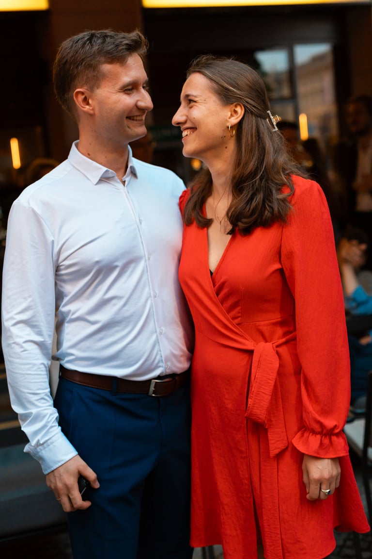A man in a light blue shirt and dark pants smiling at a woman wearing a red wrap dress at an indoor event