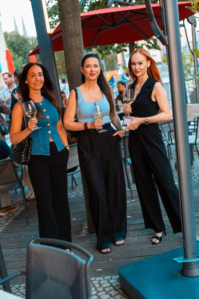 Three women holding drinks at an outdoor venue under red umbrellas and metal canopy structures