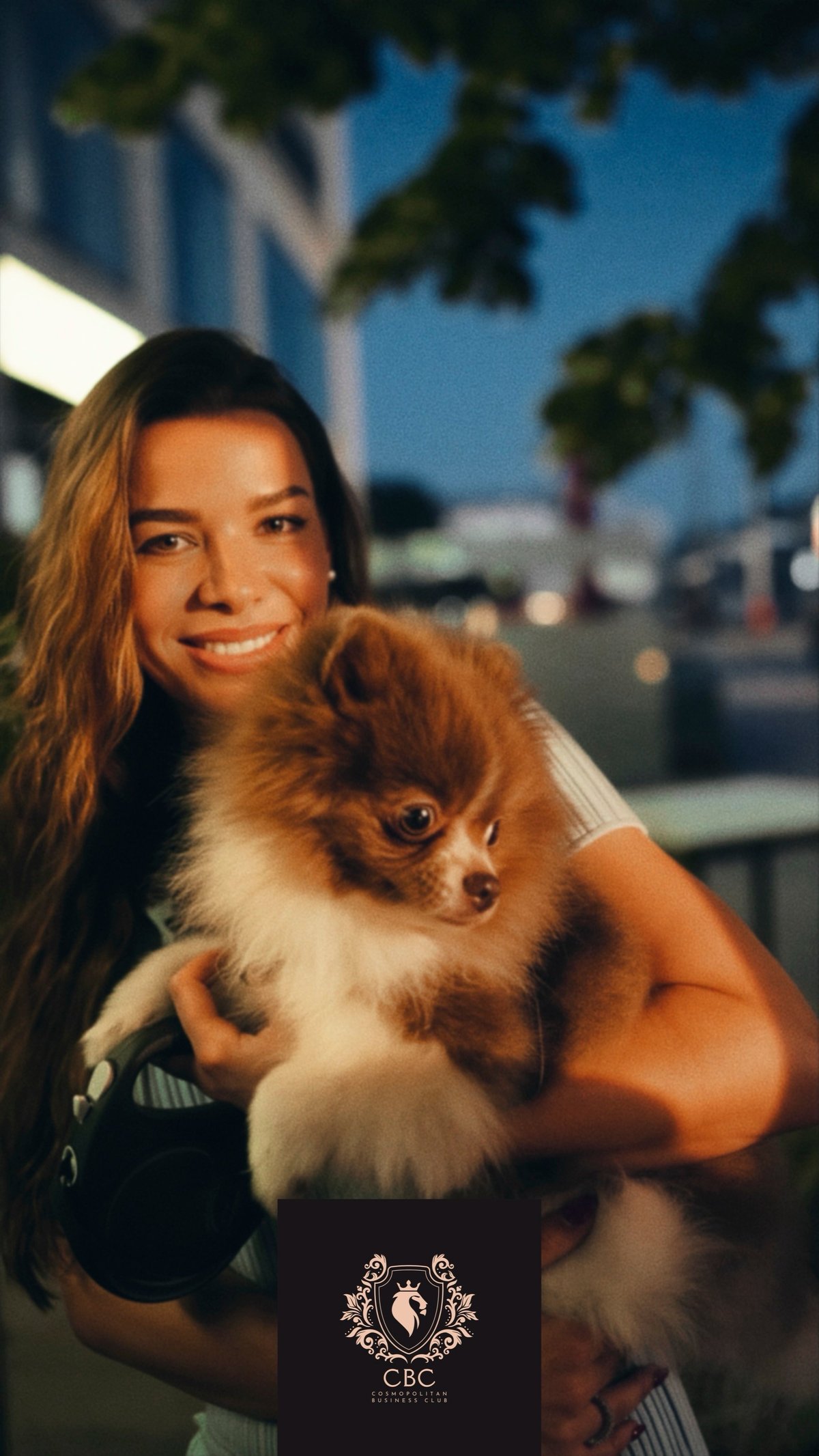 Woman smiling at camera holding brown and white Chihuahua with urban cityscape background at dusk