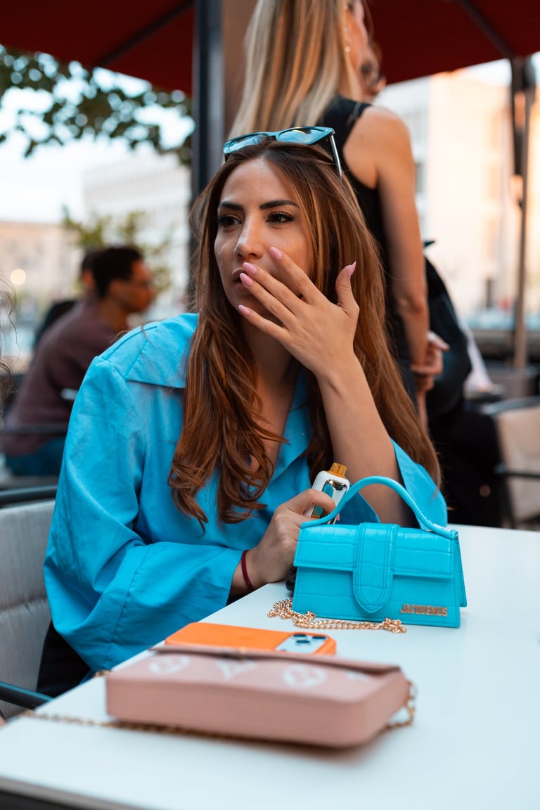 Woman in bright blue shirt sitting at outdoor cafe with turquoise handbag, sunglasses on head, hand near mouth, looking thoughtful