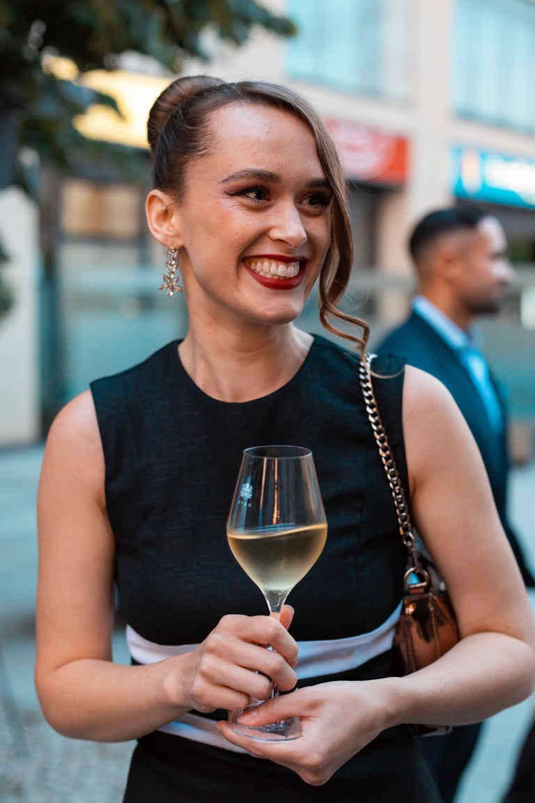 Woman smiling at outdoor event holding white wine glass, wearing black sleeveless top and brown shoulder bag