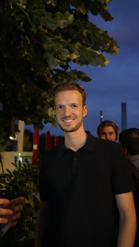 Smiling man in black polo shirt at evening outdoor event with hanging foliage and city lights in background