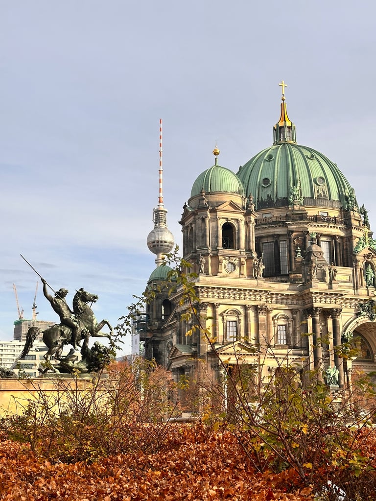 Historic cathedral with green copper dome and golden spire, a statue in foreground, surrounded by autumn foliage and city skyline