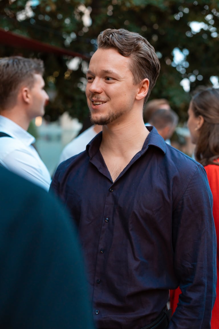 Man in dark navy button-up shirt smiling outdoors at a social gathering with other people in background