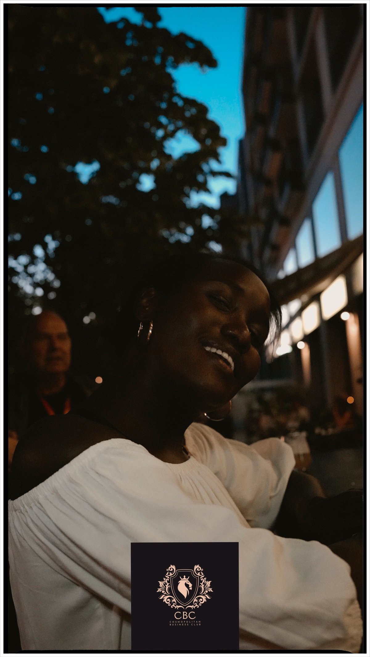Person wearing white shirt with black logo smiling at night with building and blue sky in background
